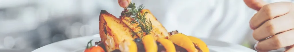 Header afbeelding voor de pagina bijbaan emmeloord. Op de foto zie je tomaten, oartjes perzik en gebakken aardappelpartjes op een wit bord liggen. Je ziet een hand een takje dille op de aardappels leggen. De achtergrond is een wit bokeh patroon.