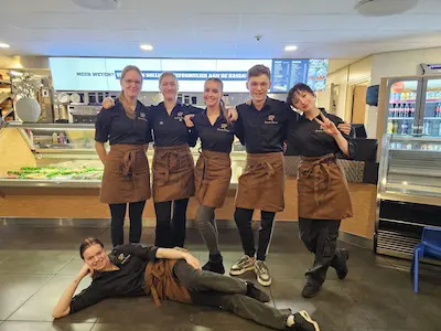 Groep collega's (tieners) die bij elkaar staan. Ze hebben hun werkkleding van Snack-Plaza aan. Ze dragen een zwarte blouse en een taupekleurige sloof.